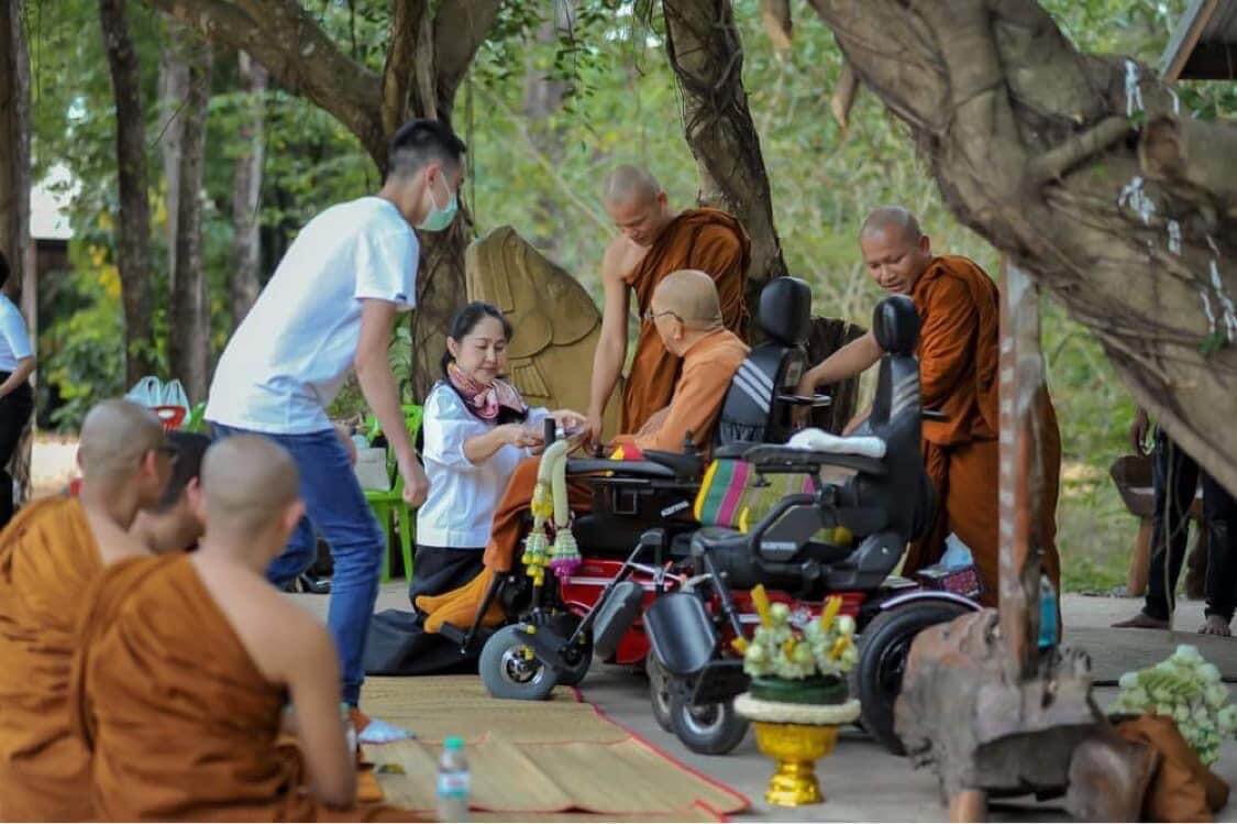 A-Thailand-monk-sitting-on-a-power-wheelchair-surrounded-by-fellow-monks-and-laypeople-offering-alms-during-a-traditional-Buddhist-ceremony-outdoors
