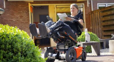 A-woman-in-a-power-wheelchair-using-tilting-and-lifting-funtion-reading-newspaper-outdoor-under-the-sunlight